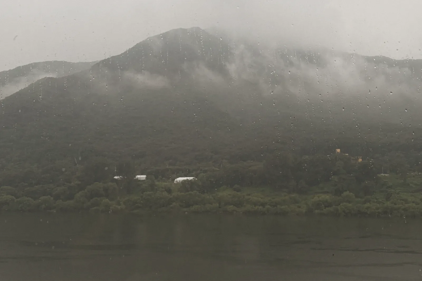 A misty, forested mountain across a river, seen through light rain, with a few small buildings at its base.