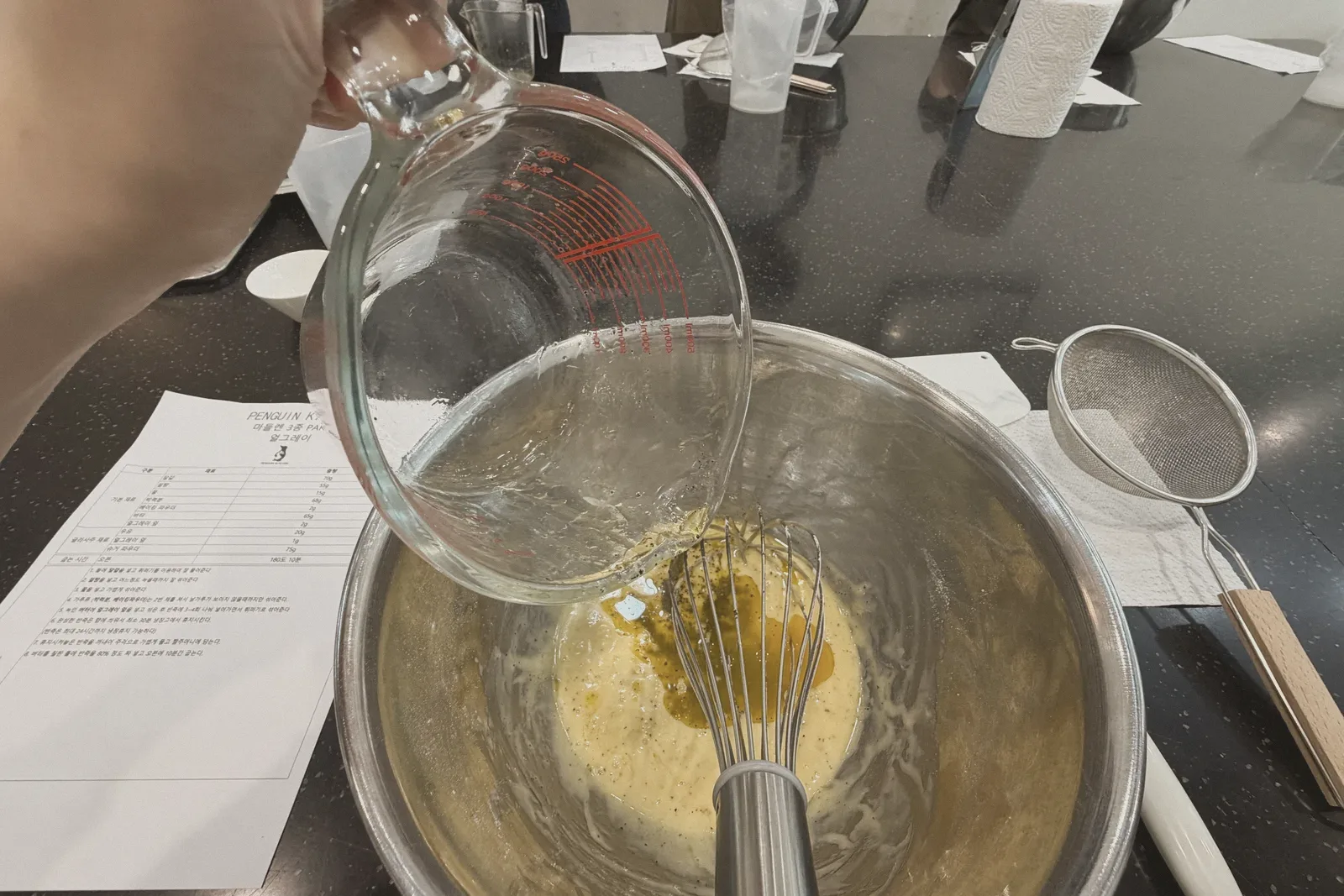 A whisk mixing a pale batter in a glass bowl, with a recipe sheet nearby.
