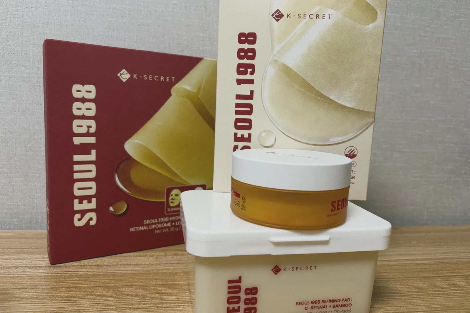 An amber-orange jar resting on a white tub of pads, in front of a maroon box and a cream box printed with sheet masks, on a wooden shelf.