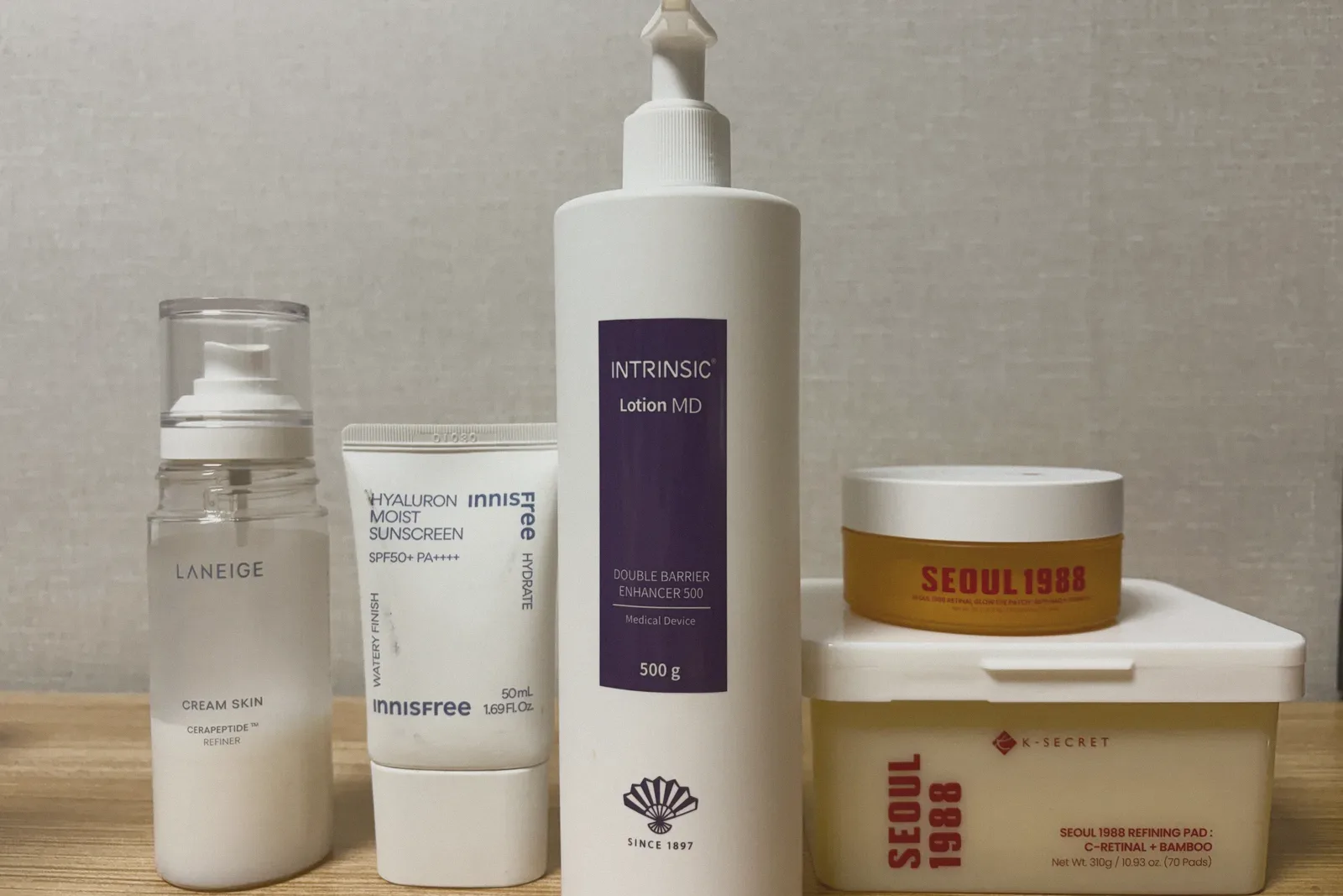 LANEIGE Cream Skin Refiner, an innisfree Hyaluron Moist Sunscreen tube, an Intrinsic Lotion MD pump bottle, and a SEOUL 1988 Refining Pad tub topped with a SEOUL 1988 Retinal Glow Eye Patch jar against a woven wallpaper background.