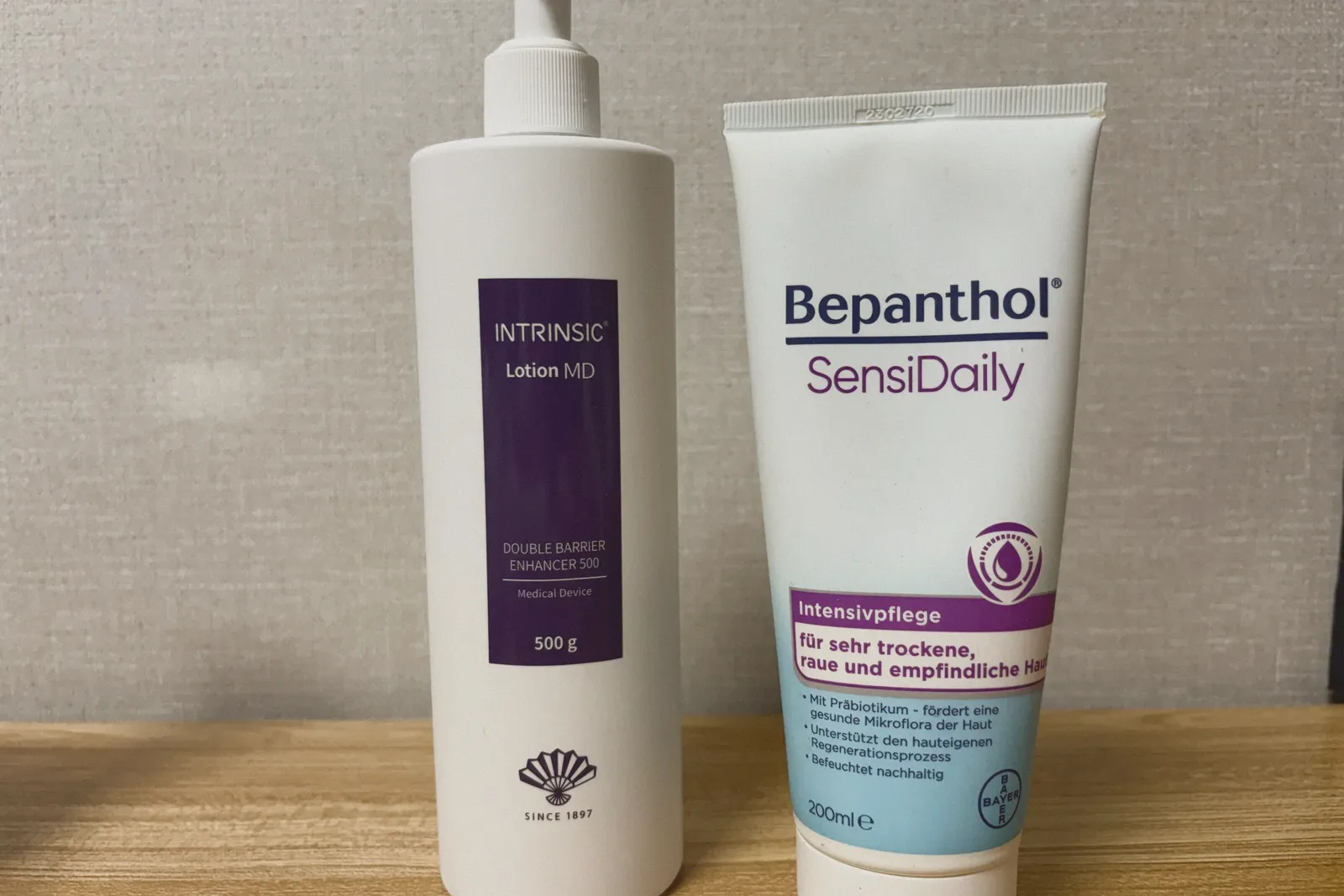 An Intrinsic Lotion MD pump bottle beside a Bepanthol SensiDaily tube against a woven wallpaper background.