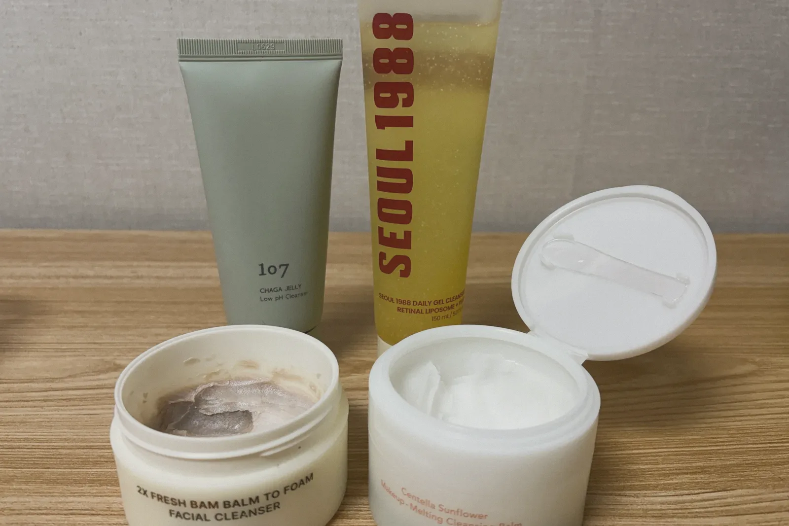 A sage-green tube and a yellow gel tube behind two open jars of cleansing balm on a wooden surface.