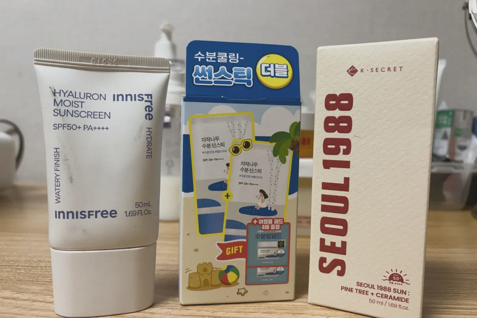 An innisfree Hyaluron Moist Sunscreen tube, a Korean sun-stick box, and a SEOUL 1988 Sun: Pine Tree + Ceramide box standing on a wooden table.